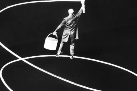 Gilbert Garcin maluje białą linię na ciemnym tle, trzymając wiadro.