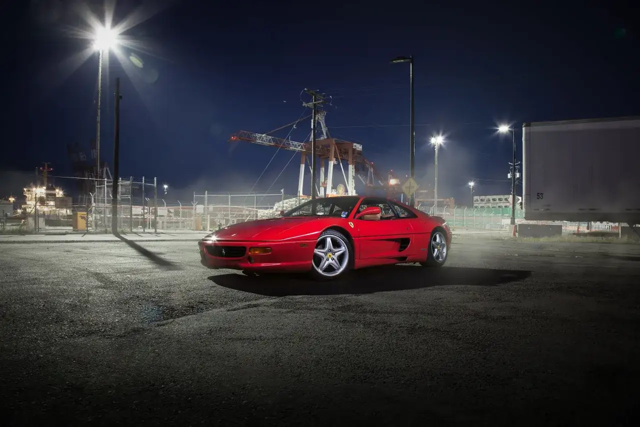 Czerwone Ferrari F355 na nocnym parkingu portowym. To wyjątkowa fotografia motoryzacyjna z widocznymi dźwigami i ciężarówką w tle.