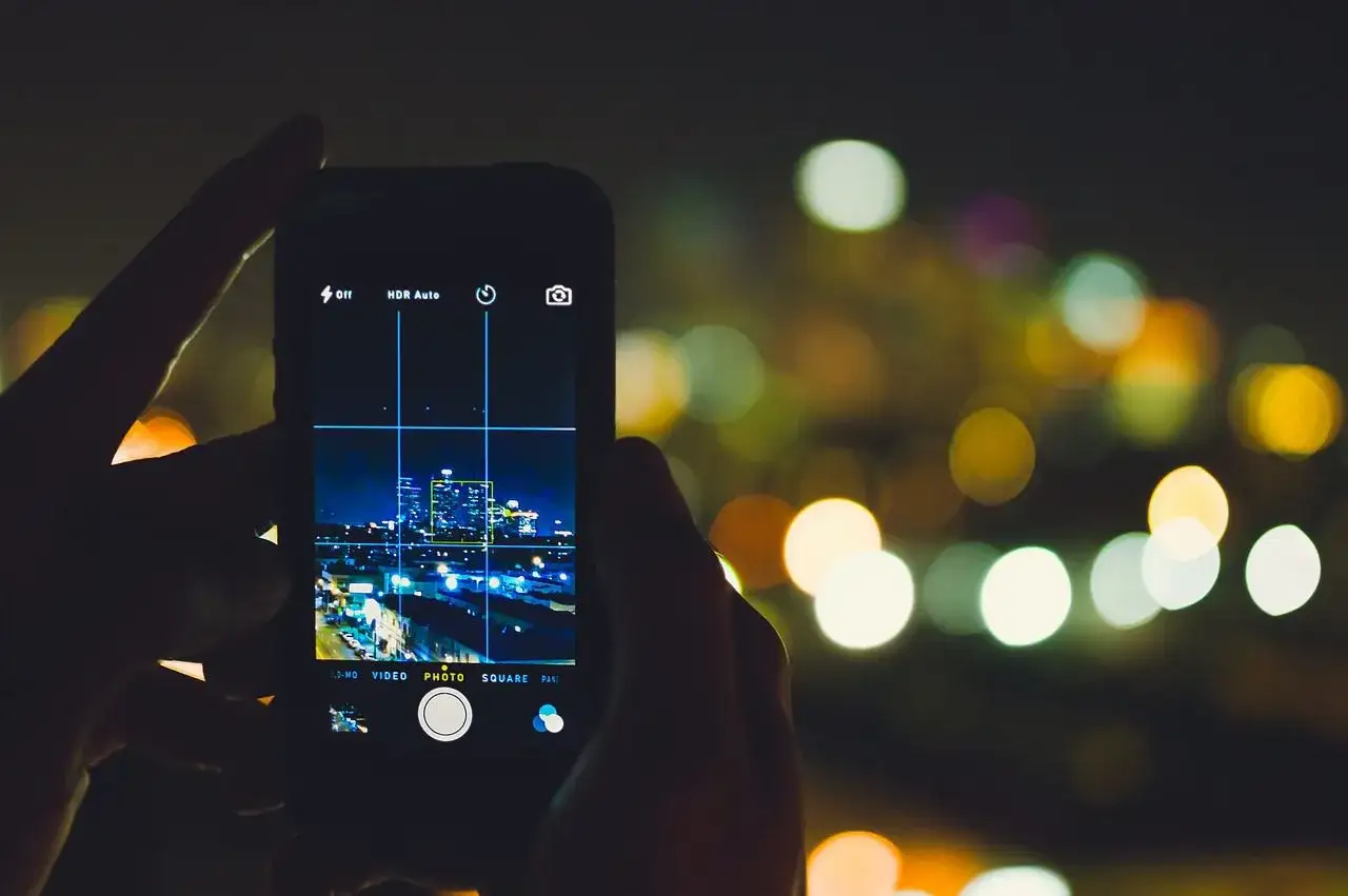 Ręce trzymają smartfon, którego aparat w telefonie rejestruje nocną panoramę miasta. Rozmyte światła tworzą magiczne tło.